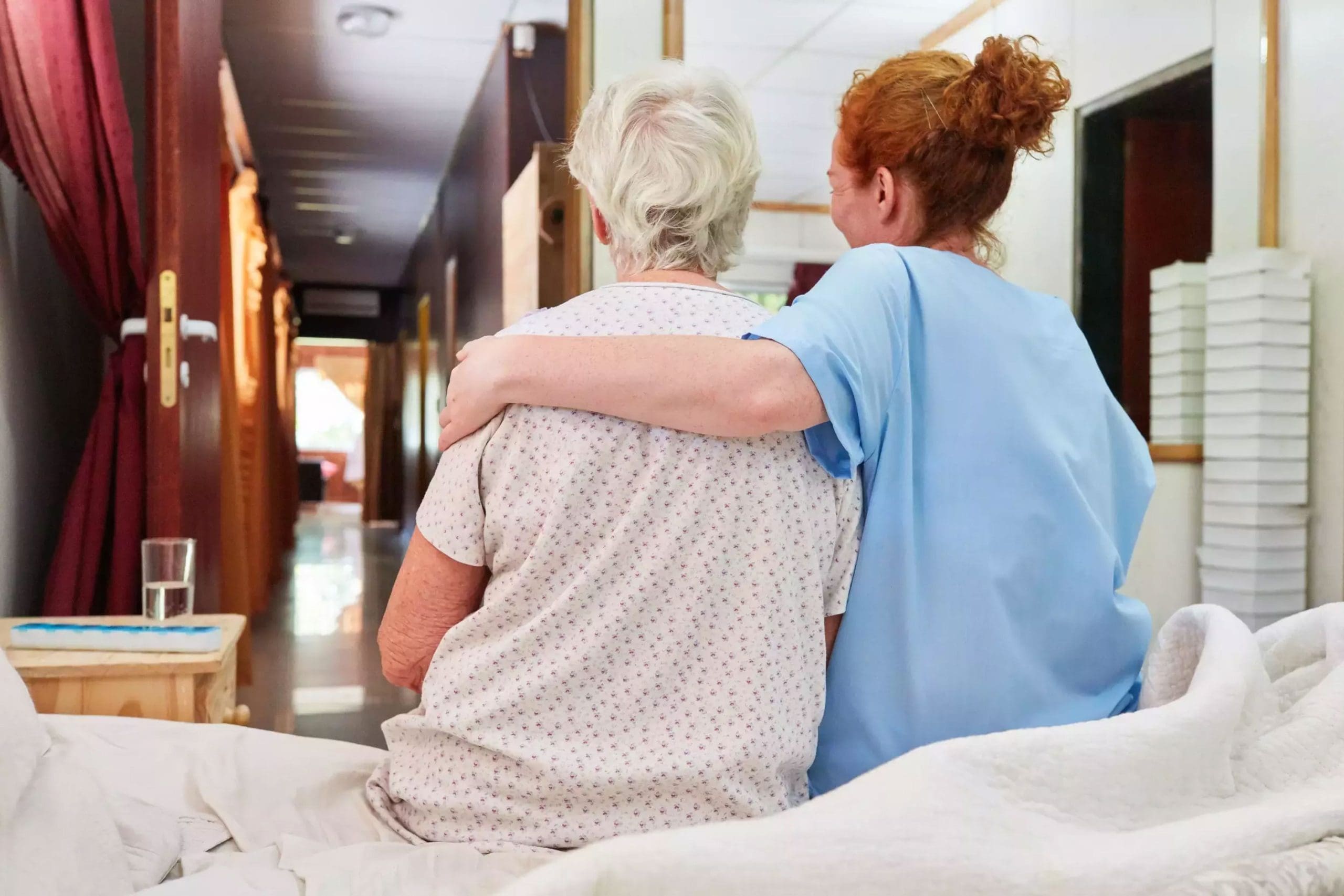 A caregiver in blue scrubs with her arm around an elderly woman sitting on a bed, offering comfort and companionship in a care home setting.