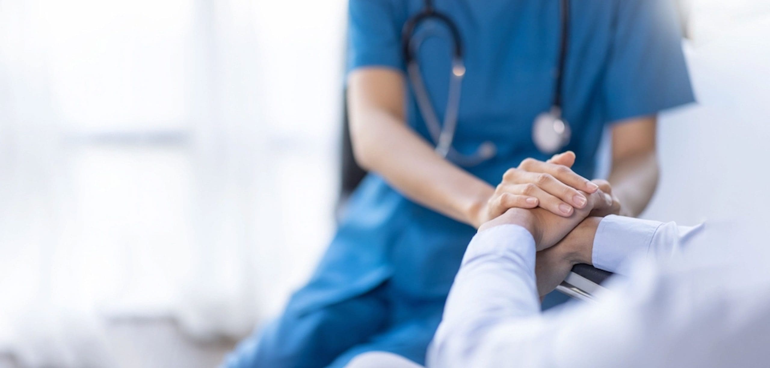 Doctor or nurse in blue scrubs gently holding a patient’s hands in a reassuring gesture in a bright medical setting.
