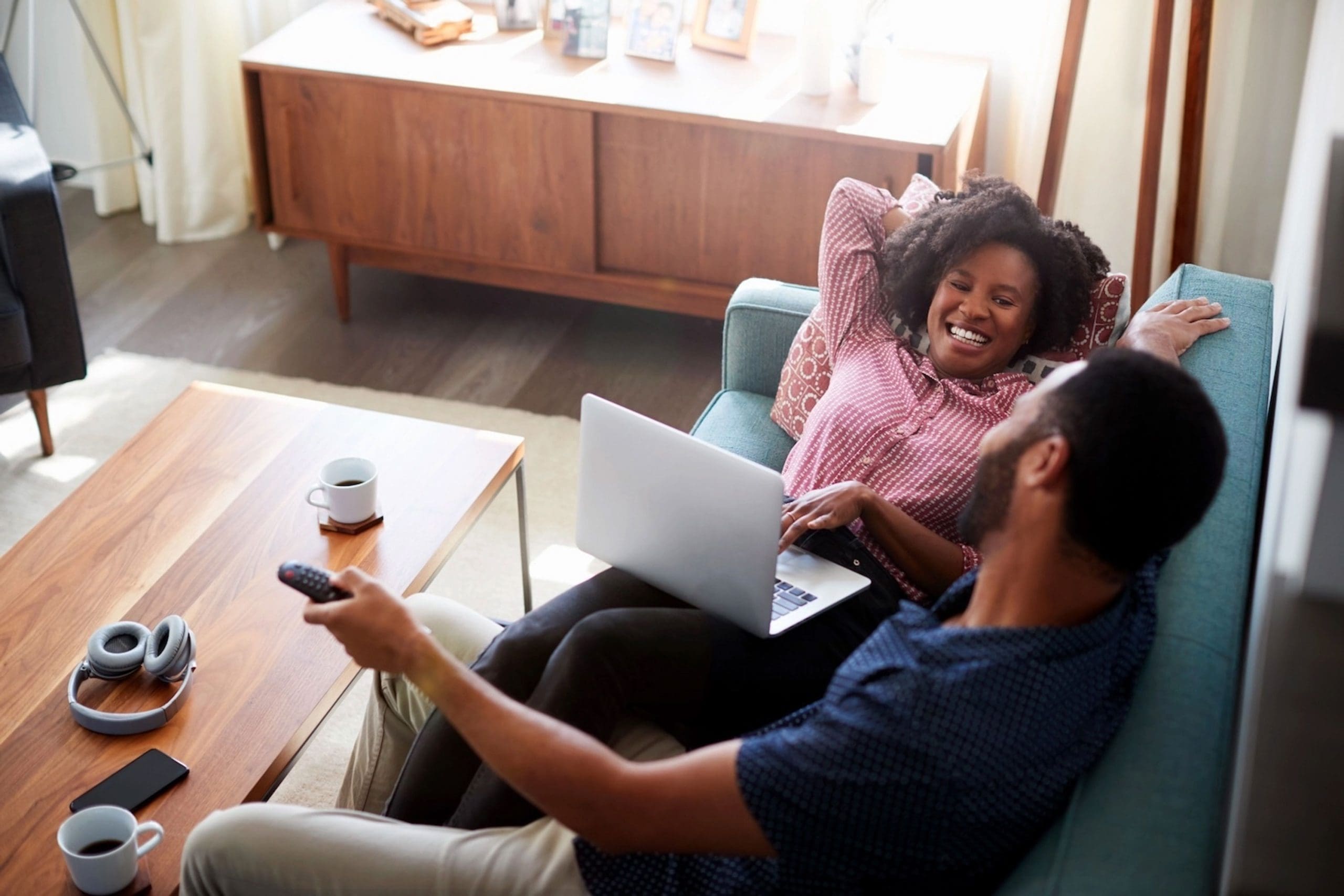 A couple relaxing on a sofa, smiling and talking while using a laptop, with coffee cups and headphones on the table in a bright living room.