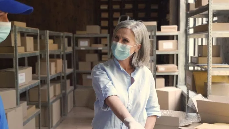 Woman wearing a face mask and gloves talking to a coworker in a warehouse filled with shelves of cardboard boxes.