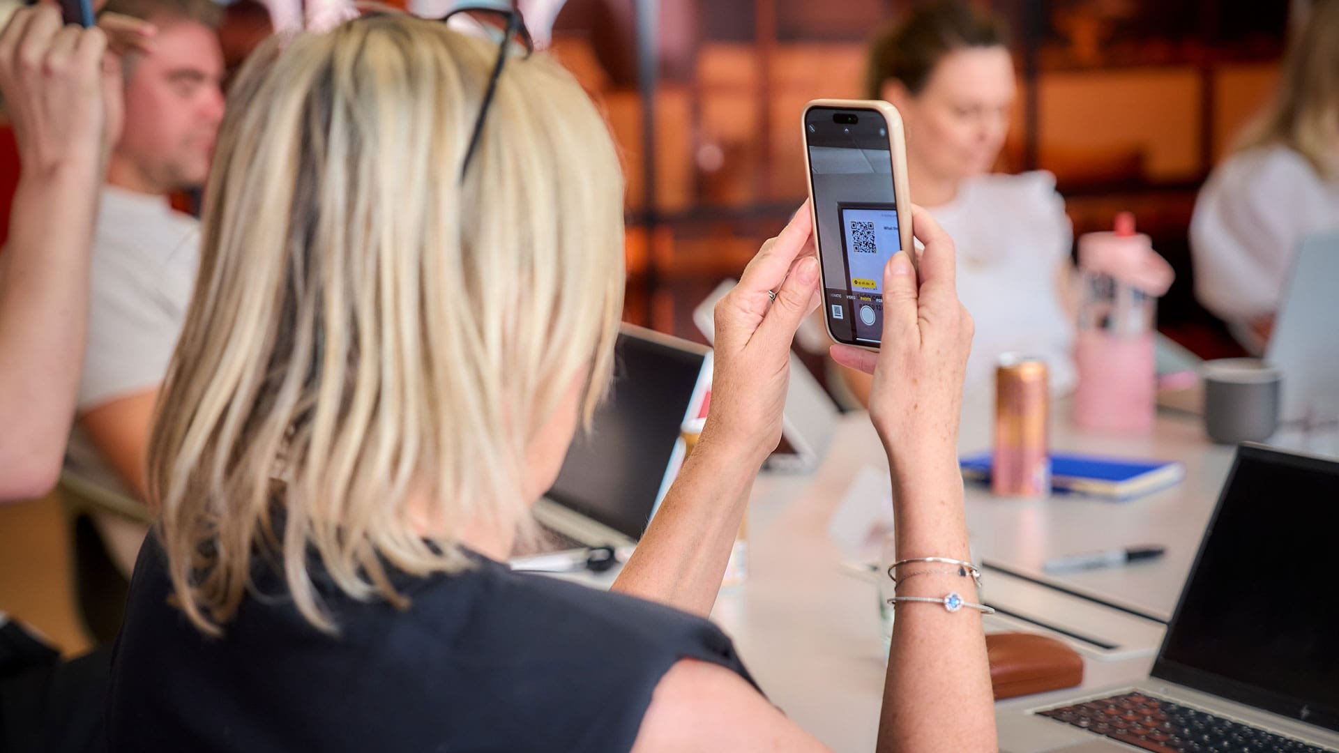 Woman with blonde hair taking a photo of a QR code on her phone during a meeting with colleagues at a table.