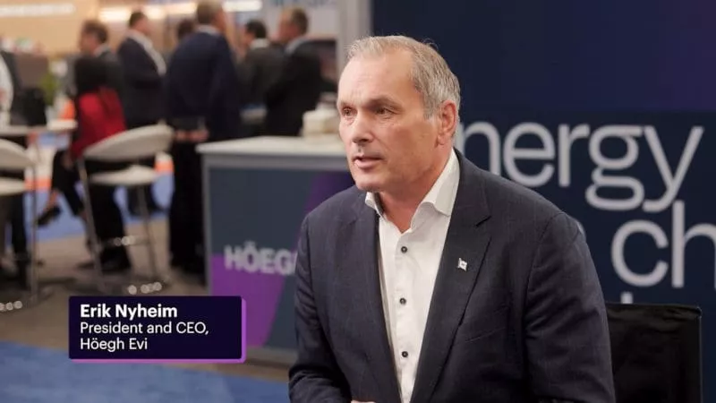 A man in a navy suit and white shirt speaks in an interview setting with a blurred conference backdrop and a caption reading “Erik Nyheim, President and CEO, Höegh Evi.”