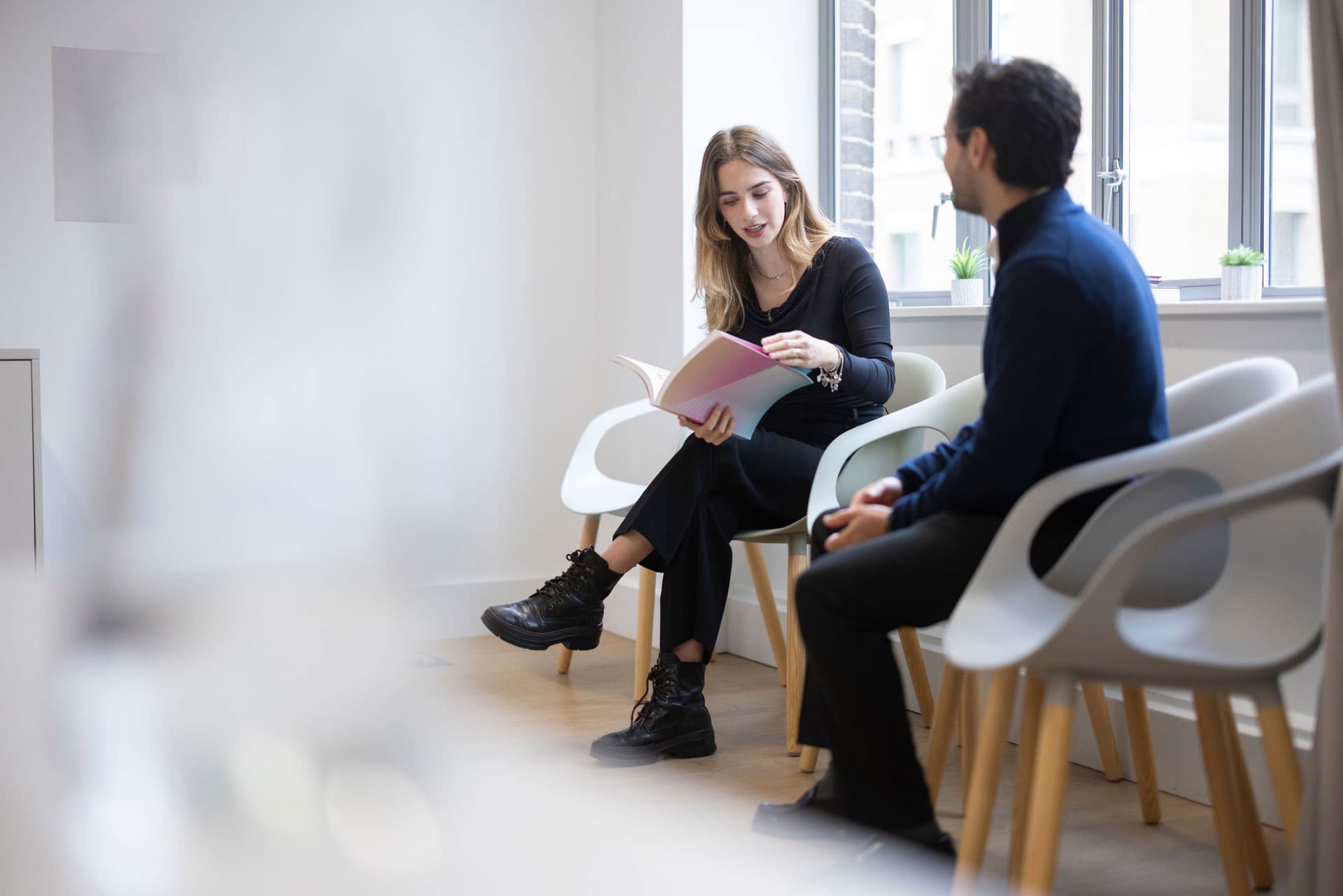 Woman showing a notebook to a man while sitting together in a bright office with modern chairs and large windows.