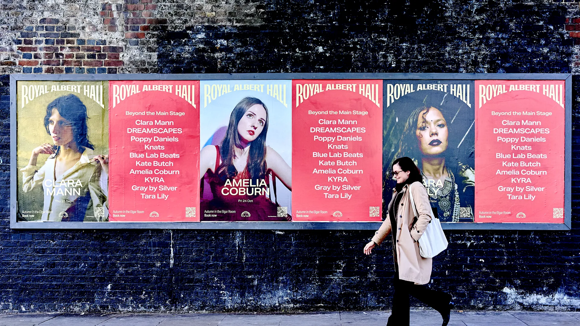 A row of six large, colorful event posters plastered on a dark brick wall along a street. Each poster has “ROYAL ALBERT HALL” at the top in bold type and features different stylized portraits of performers on vibrant red, blue, and yellow backgrounds, with event names and supporting acts listed below. A person wearing a long beige coat and carrying a white tote bag walks past in front of the posters on the sidewalk