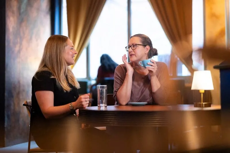 Two people sitting at a café table having a conversation, one holding a blue coffee cup.
