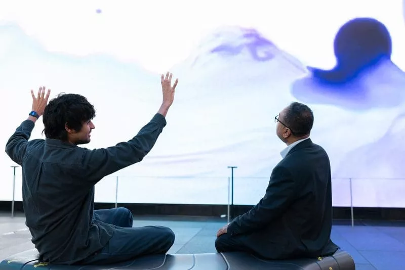 Two people sitting and talking in front of a large abstract digital screen, one gesturing with raised hands.
