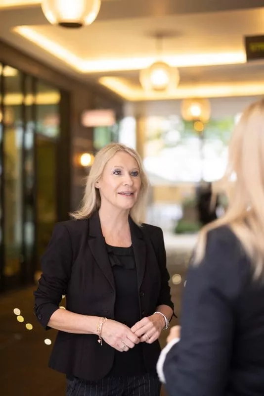 Person in a black outfit speaking with another person indoors under warm lighting.