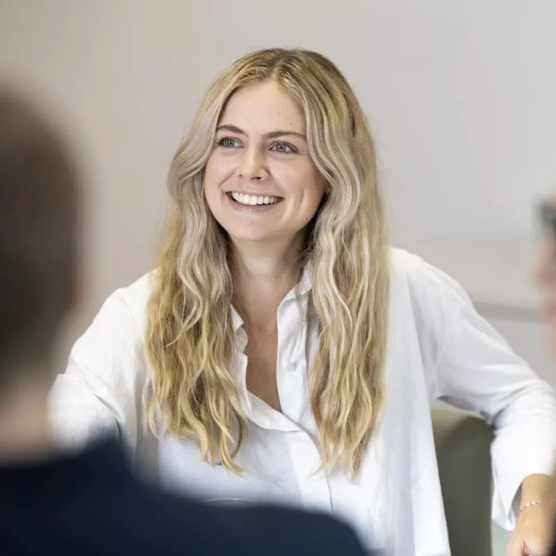 Person smiling and engaged in conversation during a meeting, wearing a white shirt.