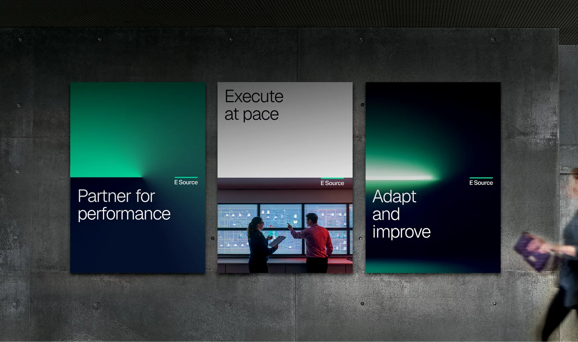 Three E Source promotional posters displayed on a concrete wall. The left poster features a green-to-navy gradient with the text "Partner for performance." The center poster shows two colleagues collaborating in front of a control room display with the headline "Execute at pace." The right poster features a dark gradient with the text "Adapt and improve." Each poster includes the E Source logo and uses a clean, modern visual style.