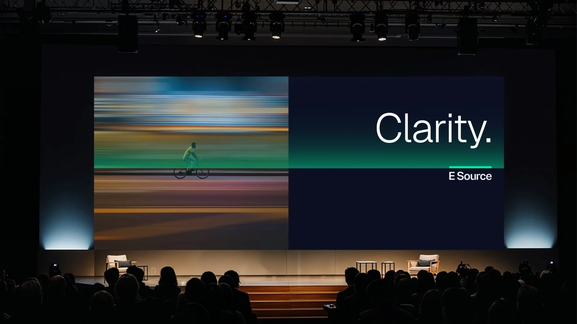 An E Source conference stage featuring a large presentation screen. The left side displays a motion-blurred image of a cyclist in transit, while the right side features the word "Clarity." in large white text above the E Source logo on a dark background with a green gradient accent. Audience silhouettes face the stage, with two armchairs and small tables set up for a speaker session.