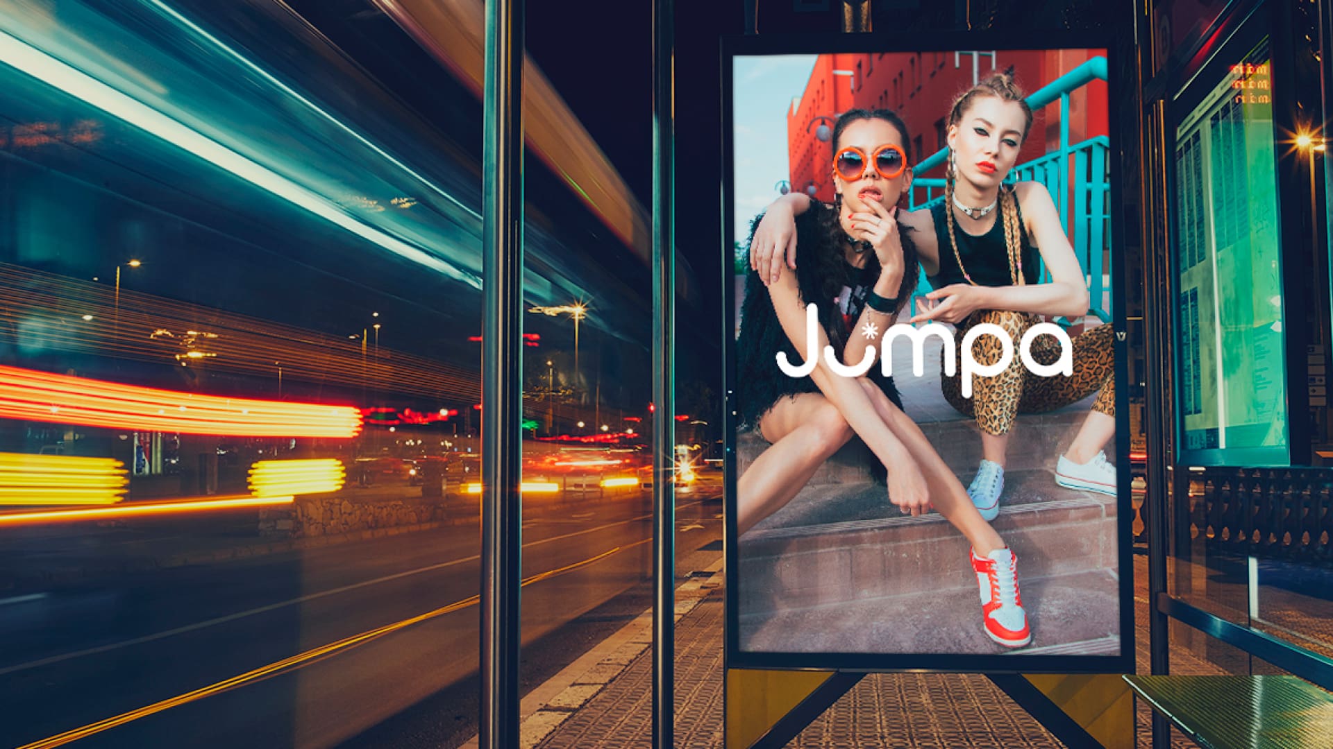 Nighttime bus stop with a brightly lit digital billboard advertising Jumpa, showing two stylish young women sitting on steps in an urban setting, with motion-blurred traffic lights streaking past on the street.