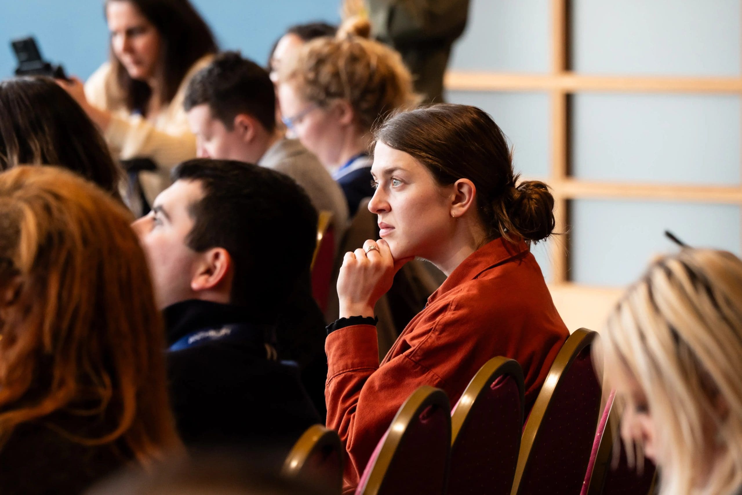 Conference attendee seated in the audience with hand resting on her chin, listening attentively during a session while other attendees sit nearby.