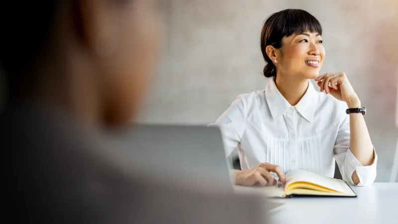 A woman in a white blouse sits at a desk with an open notebook, smiling and looking off to the side during a meeting or conversation. A blurred coworker and laptop are visible in the foreground, with soft natural light brightening the modern office setting.