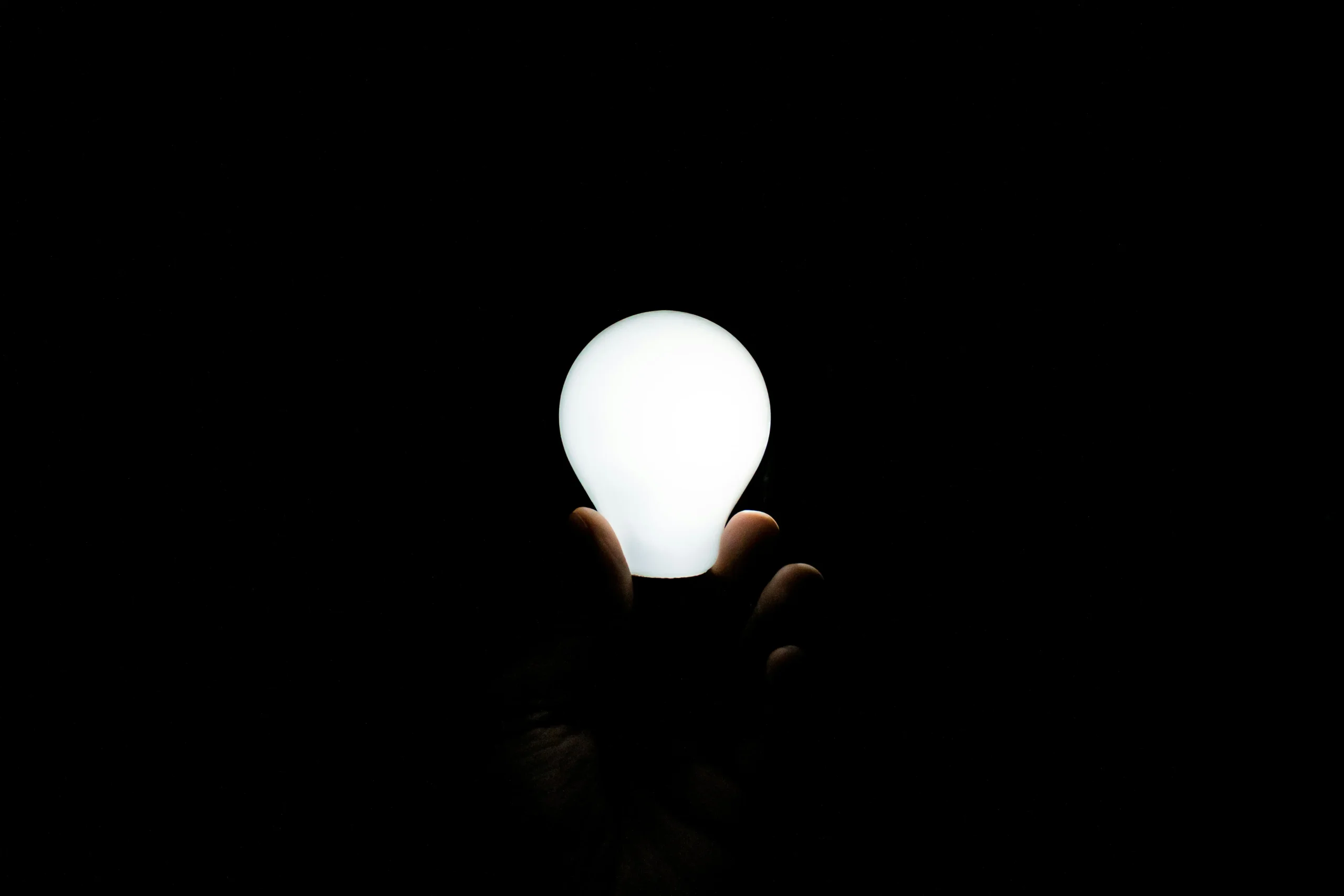A glowing white light bulb is held upright in a person's hand against a completely black background. The bulb illuminates the fingertips beneath it, creating a stark contrast between the bright light and the surrounding darkness.
