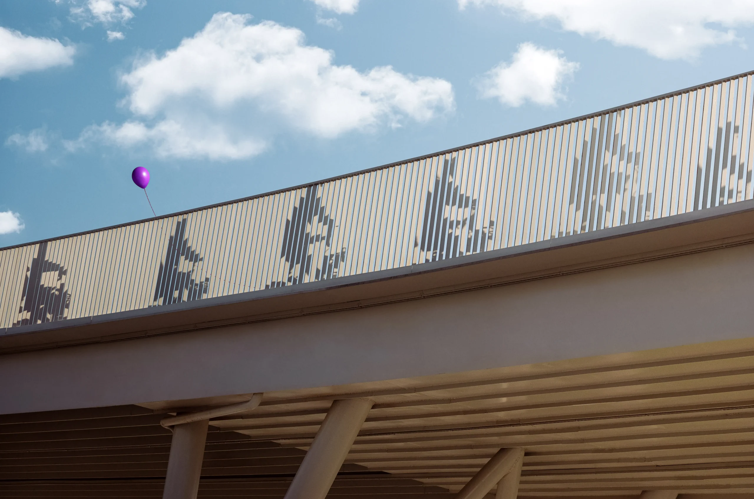 Shadows of several people are cast onto the vertical slats of a pedestrian bridge railing, creating silhouette-like figures against a bright sky with scattered clouds. A single purple balloon floats above the railing near the left side of the image. The bridge structure and support columns are visible below, viewed from a low angle.