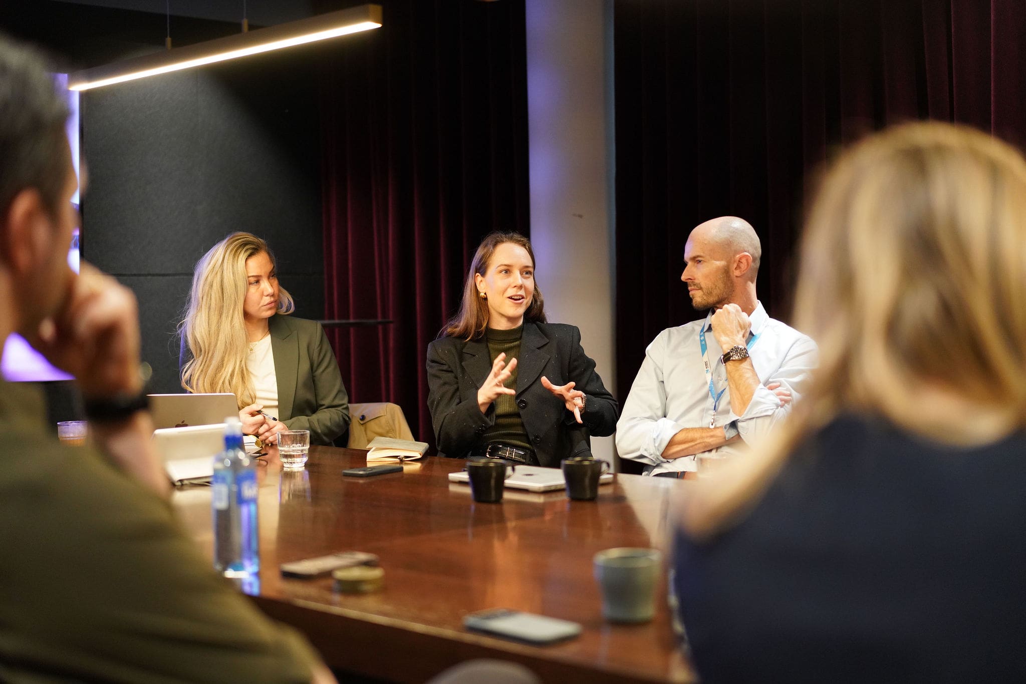 A group of professionals sits around a conference table during a Brand Rebels event. A woman in the center gestures as she speaks while two colleagues listen attentively. A laptop, notebooks, coffee cups, water bottles, and smartphones are on the table, with other attendees blurred in the foreground, creating a candid meeting atmosphere.