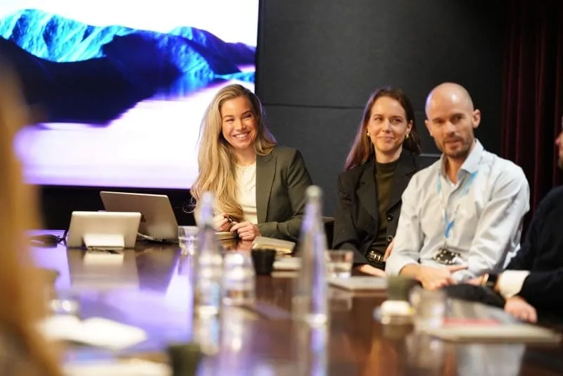 Three professionals sit around a conference table during a Brand Rebels event. A smiling woman with a laptop is speaking while two colleagues listen beside her. Water bottles, notebooks, and glasses are on the table, and a large screen displaying a blue-toned mountain landscape is visible in the background. The foreground is softly blurred, emphasizing the discussion.