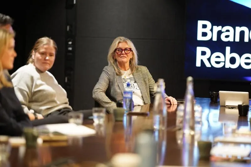 Participants sit around a conference table during a Brand Rebels Brandpie event, listening attentively to a speaker off camera. A woman in a grey blazer and red-framed glasses is in focus, with a presentation screen visible in the background. Water bottles, coffee cups, notebooks, and a laptop are arranged on the table, creating a professional meeting setting.