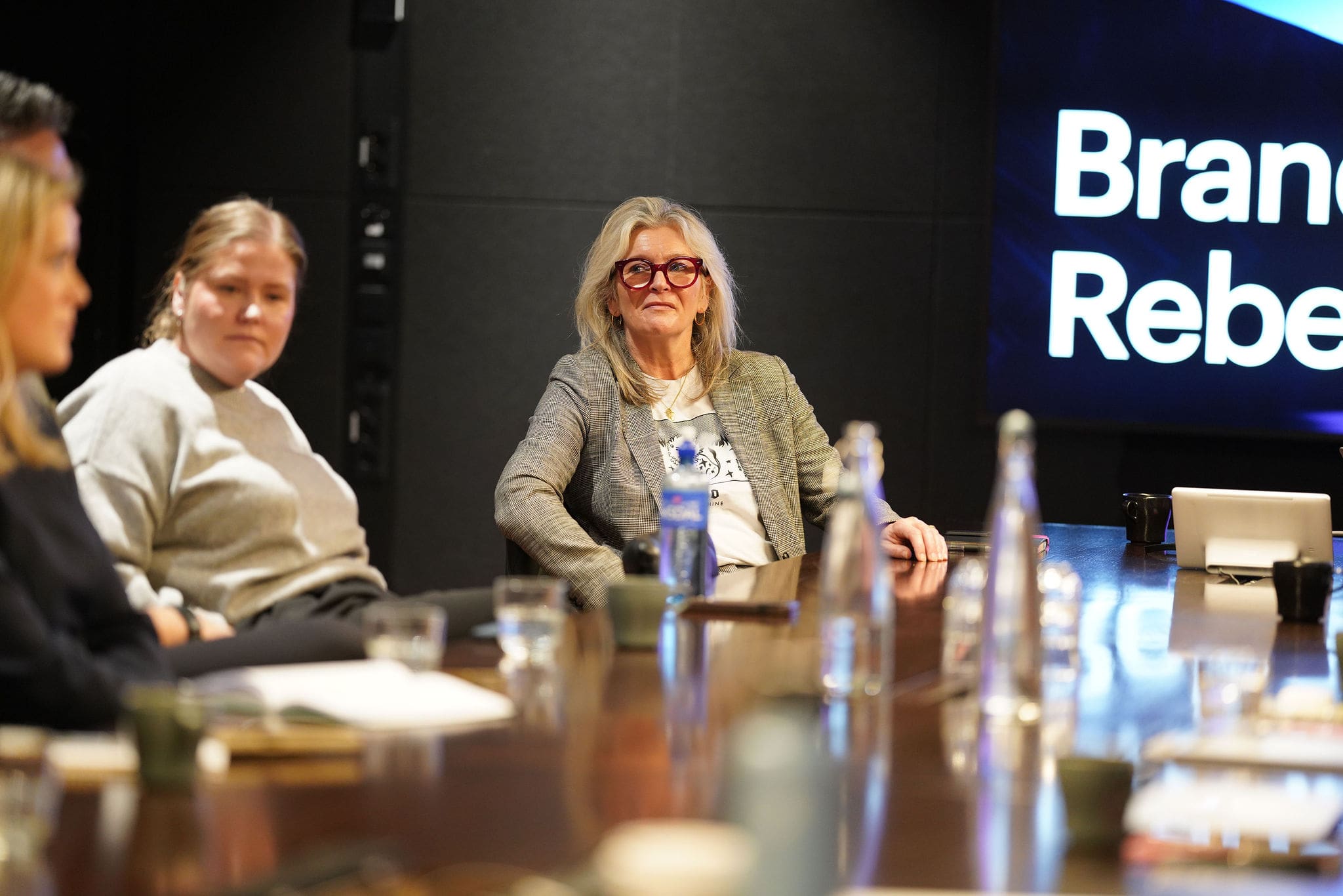 Participants sit around a conference table during a Brand Rebels Brandpie event, listening attentively to a speaker off camera. A woman in a grey blazer and red-framed glasses is in focus, with a presentation screen visible in the background. Water bottles, coffee cups, notebooks, and a laptop are arranged on the table, creating a professional meeting setting.