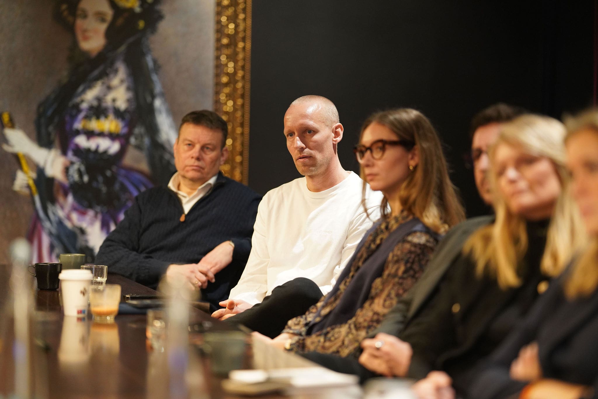 Attendees sit around a conference table listening attentively during a Brand Rebels Brandpie event. A man in a white sweater is in sharp focus at the center, while other participants are softly blurred in the foreground and background. Coffee cups, water glasses, and notebooks line the table, and a large framed portrait is visible on the wall behind the group.