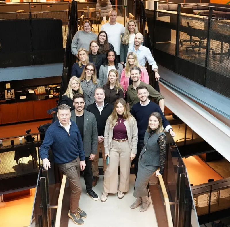 The Brand Rebels team poses together on a staircase in a modern office, smiling toward the camera. The group is arranged across several steps, showcasing a diverse mix of colleagues in a bright, contemporary workspace with open meeting areas and industrial-style design.