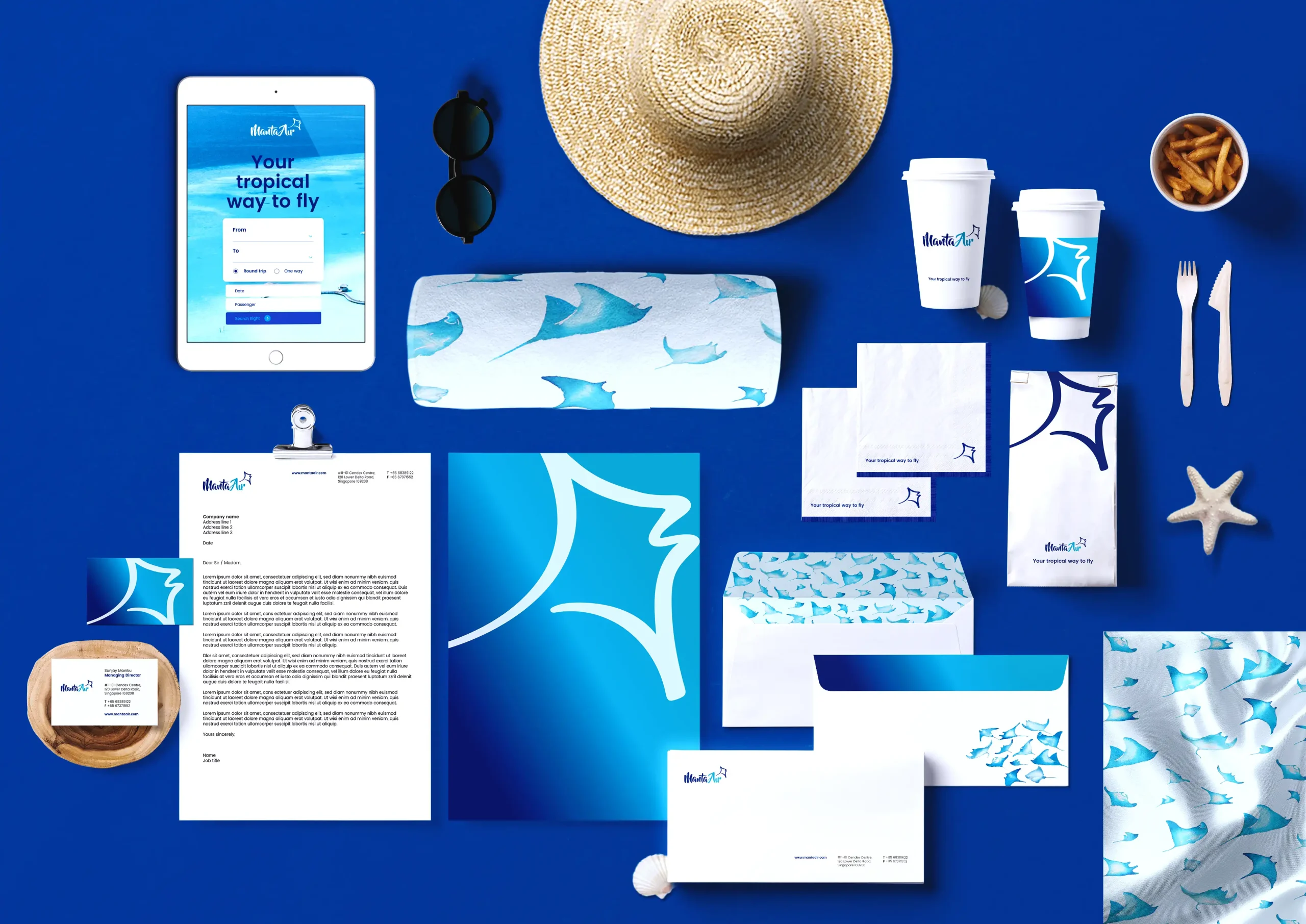 A flat lay of Manta Ait branded materials arranged on a bright blue background, including a tablet displaying a flight booking page, stationery, envelopes, business cards, branded coffee cups, tissue paper, packaging, sunglasses, a straw hat, cutlery, fries, and a starfish. The cohesive blue-and-white branding features tropical-inspired patterns and the airline's logo throughout.