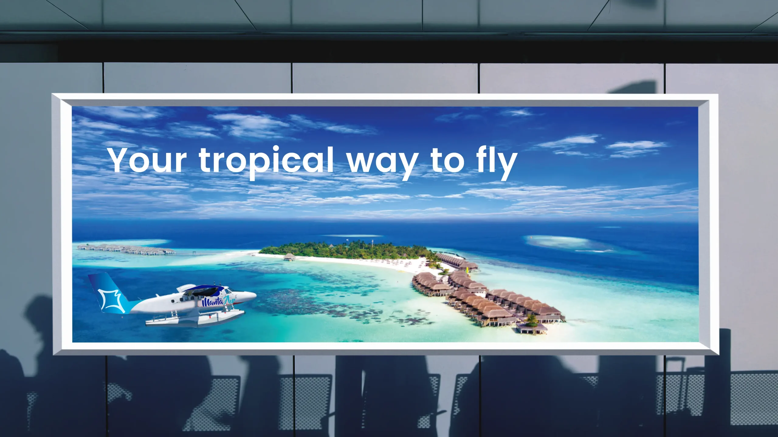 A large airport billboard advertisement featuring an aerial view of a tropical island with turquoise water and overwater villas. A Manta Air seaplane flies across the foreground beneath the slogan, "Your tropical way to fly," promoting travel to island destinations. Silhouettes of travelers in an airport terminal appear below the illuminated display.