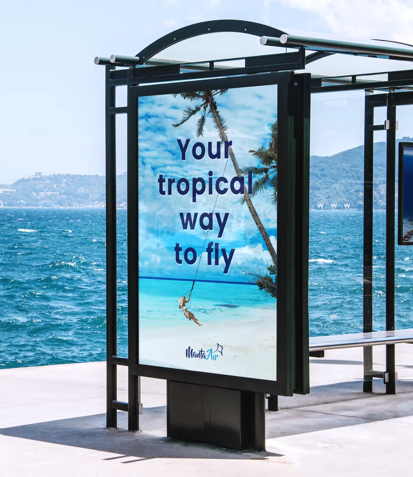 A bus stop advertisement featuring a tropical beach scene with a palm tree and a person swinging over white sand. Large text reads, "Your tropical way to fly," with the Manta Air logo at the bottom. The bus shelter overlooks a blue ocean with distant hills under a bright sky.