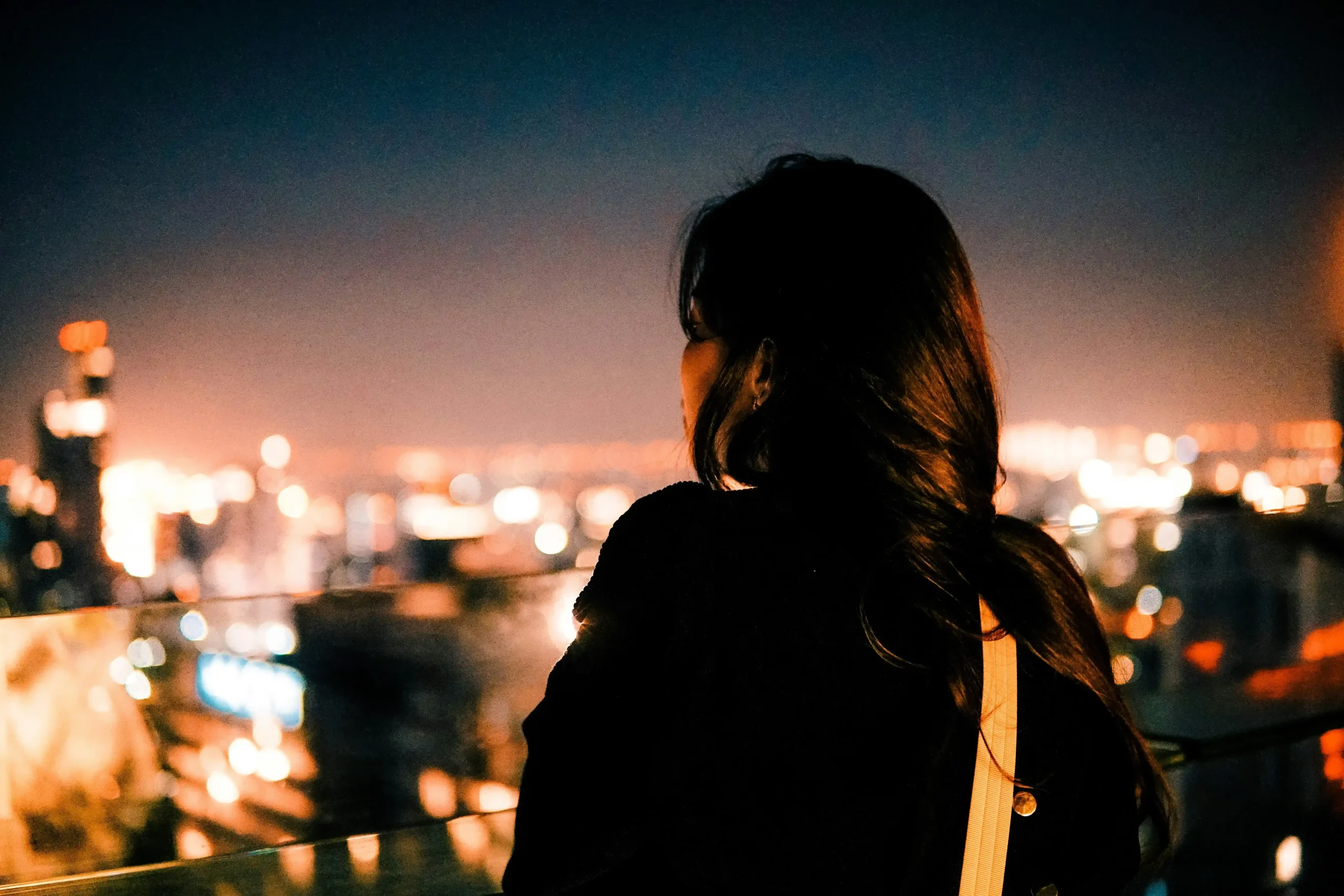 Three women having a discussion in a modern office or coworking space, with one woman listening attentively across a laptop while another gestures in the foreground; potted plants line a shelving unit in the background. A woman seen from behind on a rooftop or balcony at night, looking out over a softly blurred cityscape filled with warm glowing lights beneath a dark blue sky.