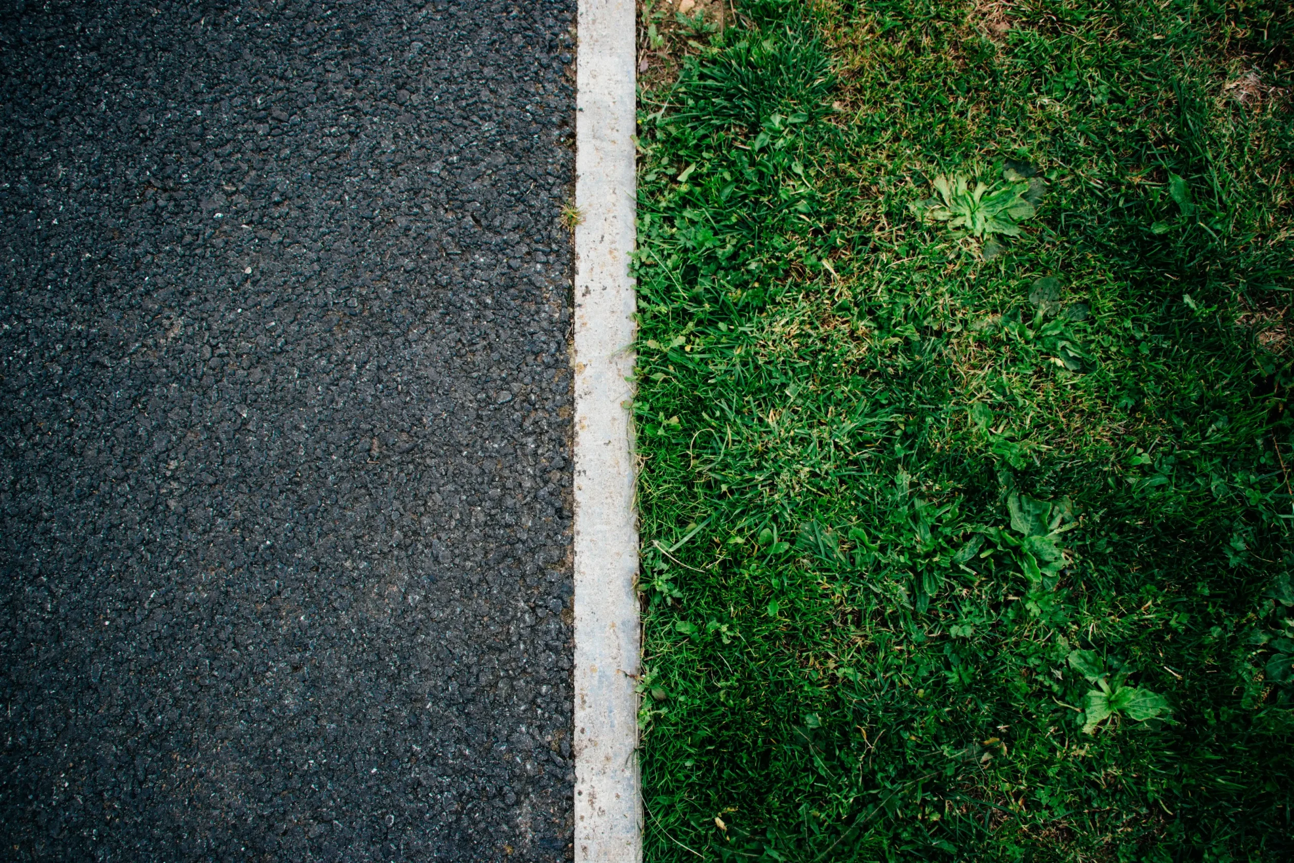Top-down view of a dark asphalt path beside a strip of green grass, separated by a narrow light-colored concrete curb.