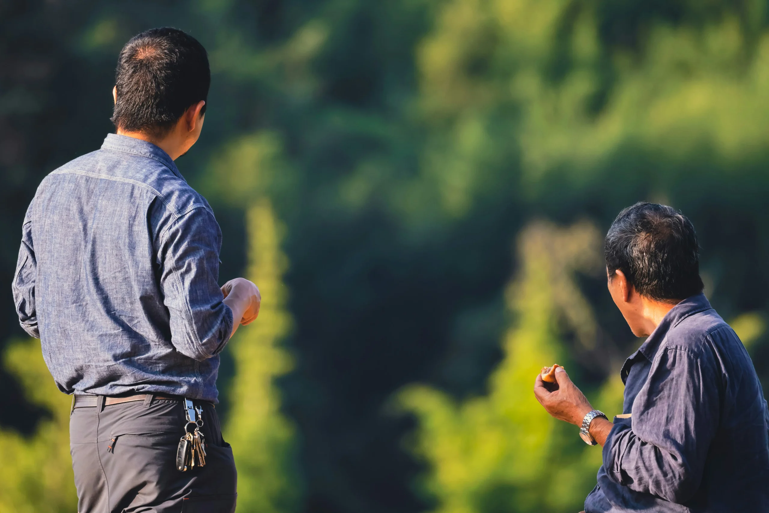 Two men outdoors in a sunlit natural setting, viewed mostly from behind; one is standing with keys clipped to his waistband, while the other is seated and holding a small piece of food, with blurred green foliage in the background.