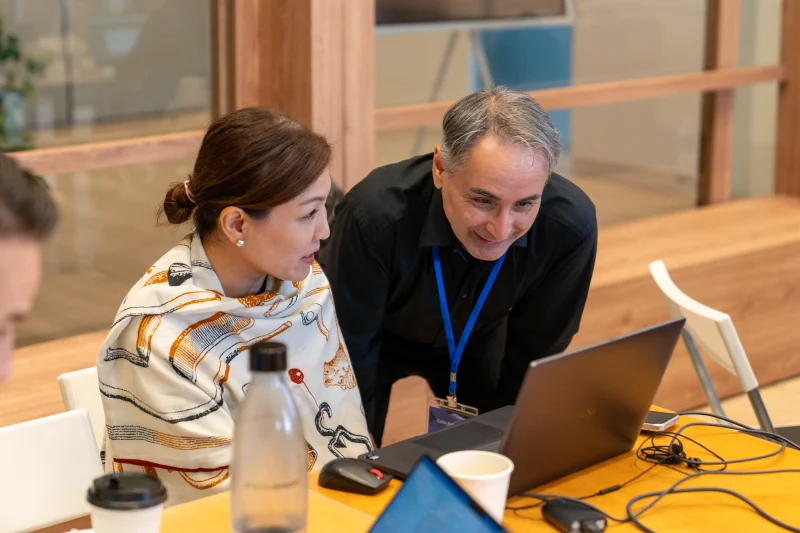 Two colleagues collaborating at a wooden table, leaning toward a laptop screen during a workshop or meeting. A woman in a patterned shawl sits beside a man wearing a black shirt and blue lanyard, with drinks, cables, and electronic devices spread across the table.