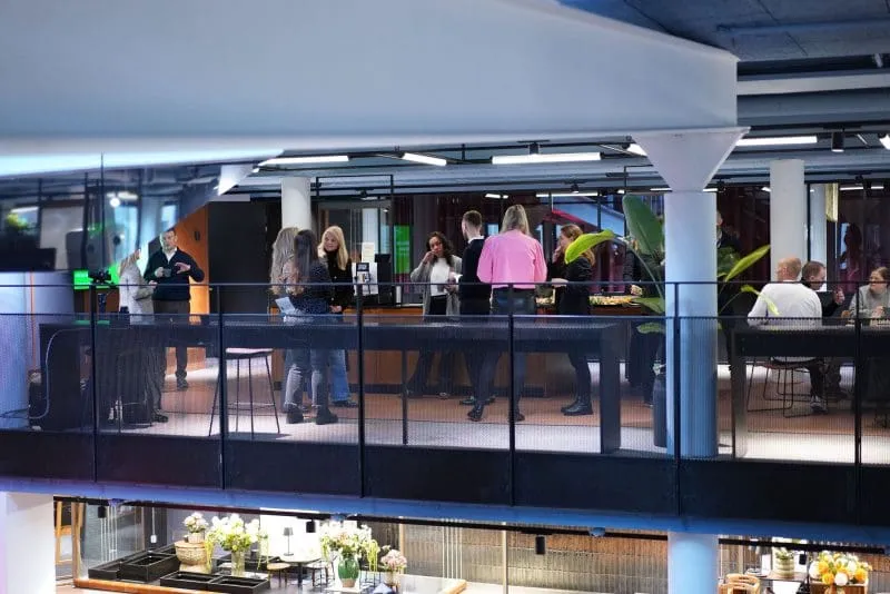 A group of people mingle and chat at an indoor networking or social event on an upper-level balcony, with refreshments set out nearby. The modern venue features exposed ceilings, glass walls, black railings, white columns, and large green plants, with a café or dining area visible below.