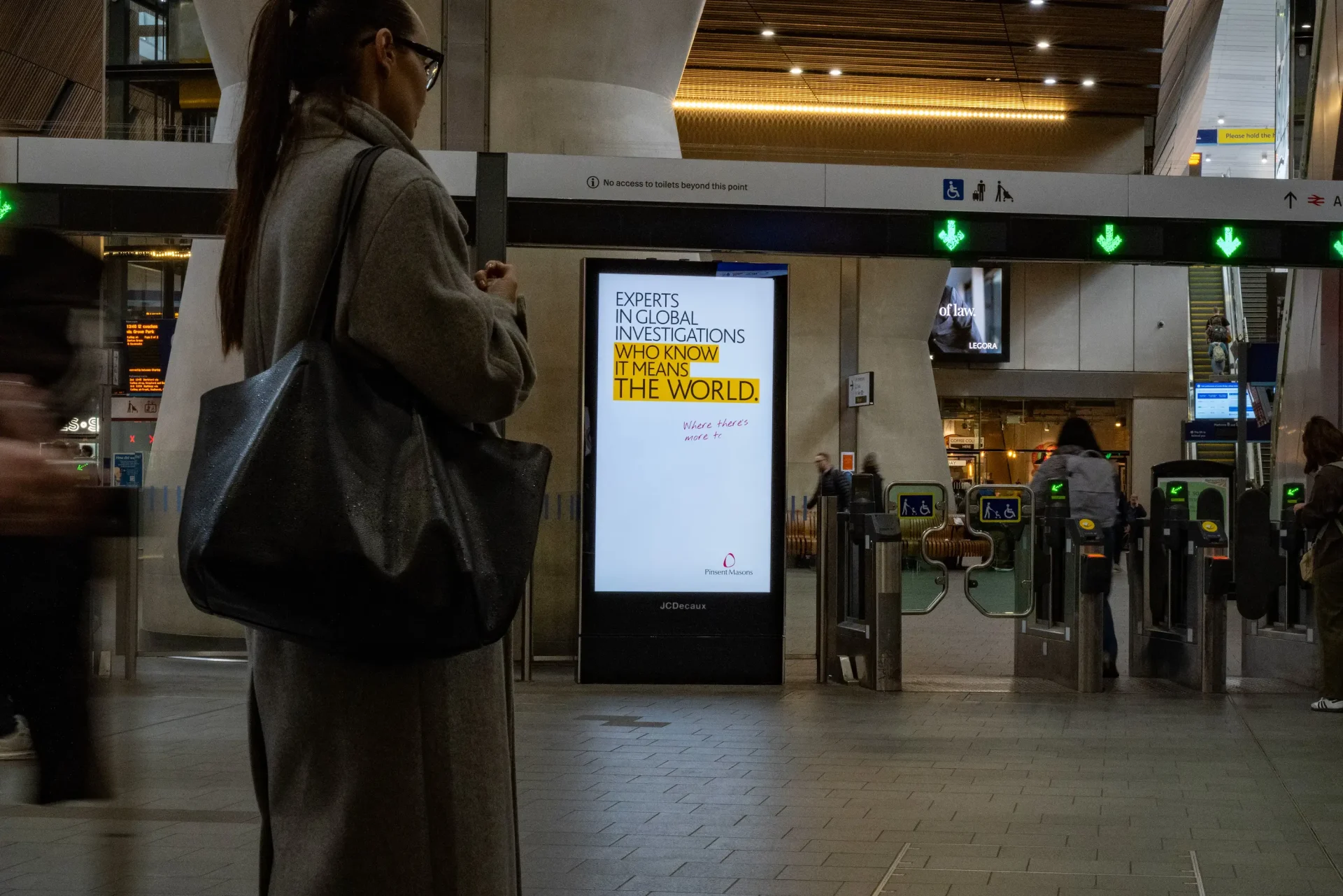 Pinsent Masons digital advert beside station ticket gates, reading “Experts in global investigations who know it means the world”, with yellow highlights.