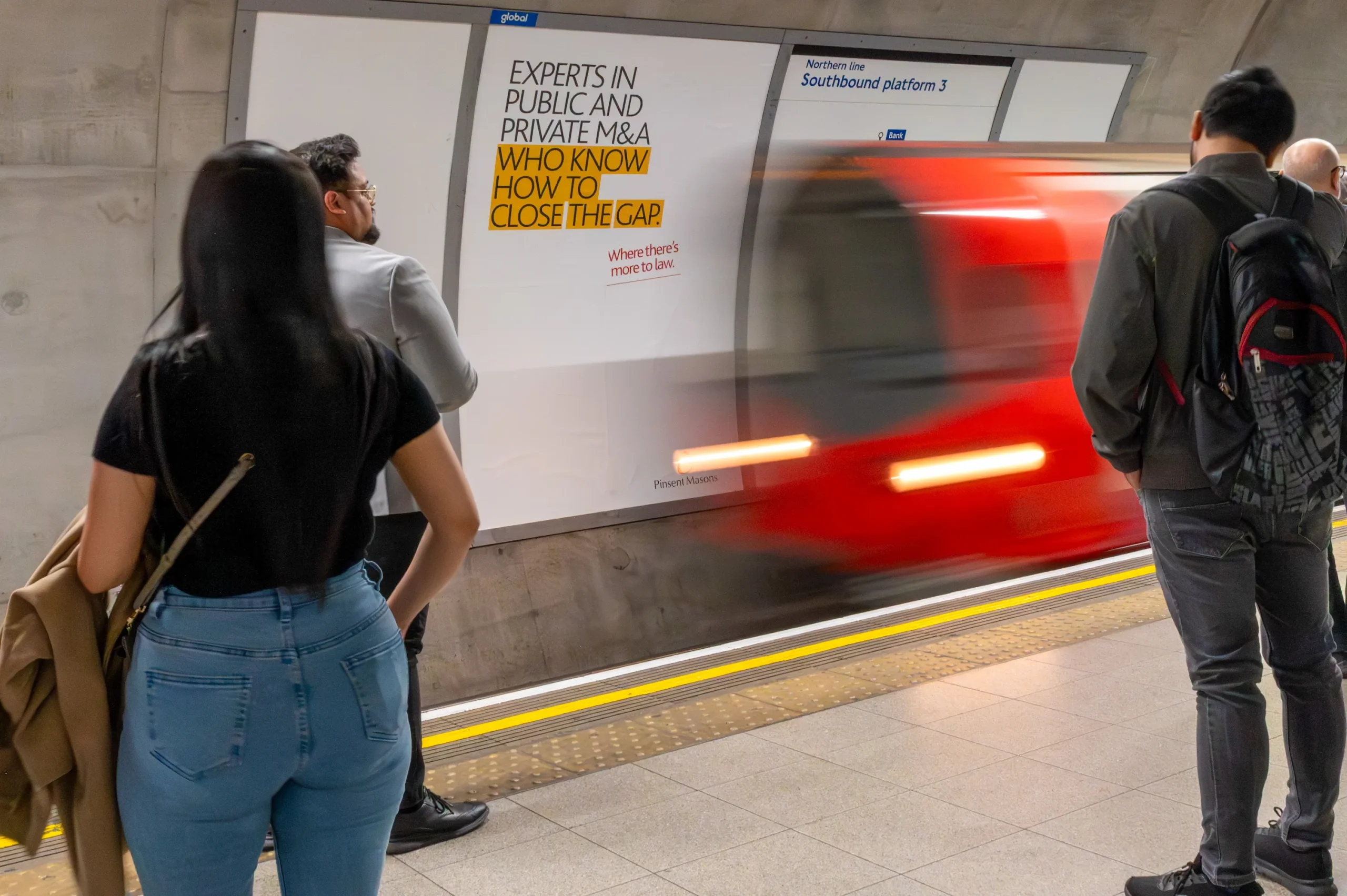Commuters and a passing Tube train at Bank station, beside a Pinsent Masons advert reading “Experts in public and private M&A who know how to close the gap.”