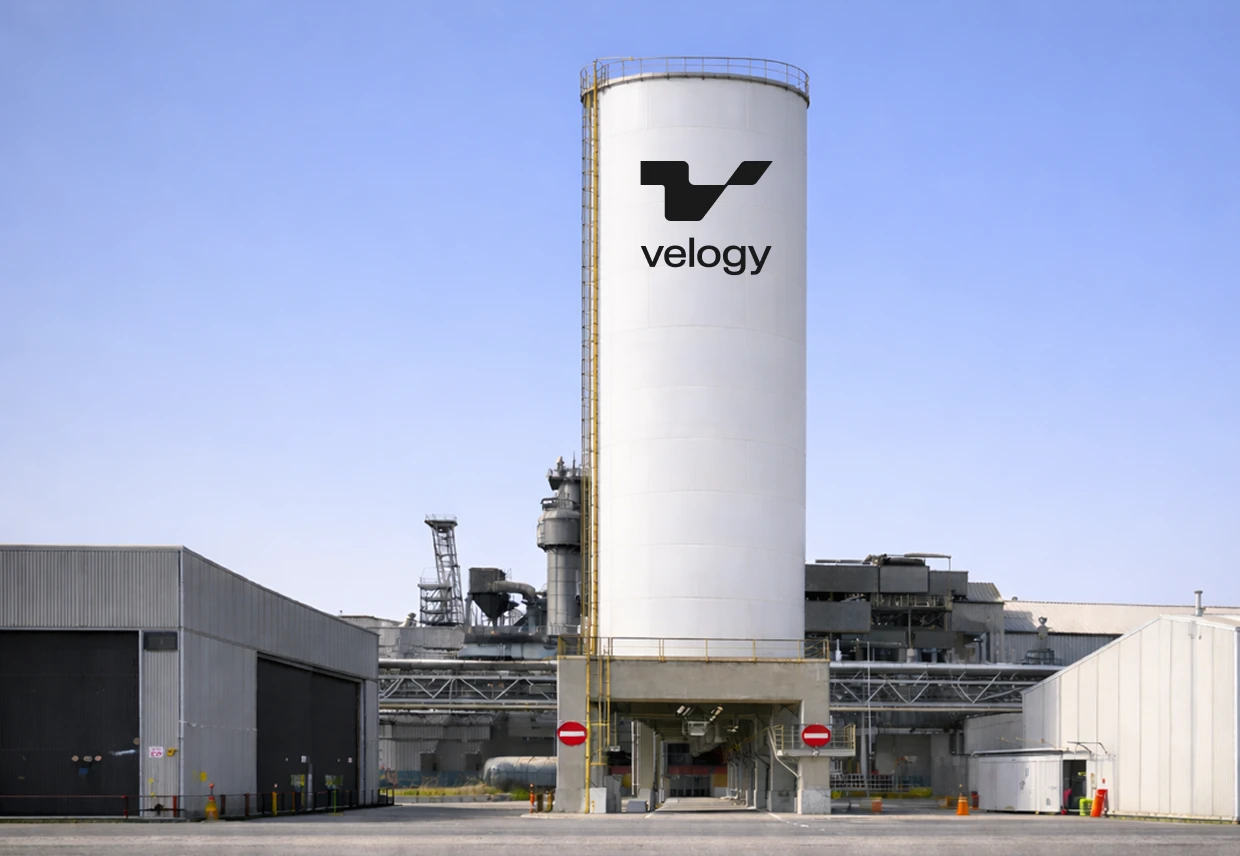 Tall white industrial silo displaying the black Velogy symbol and wordmark above surrounding factory buildings.