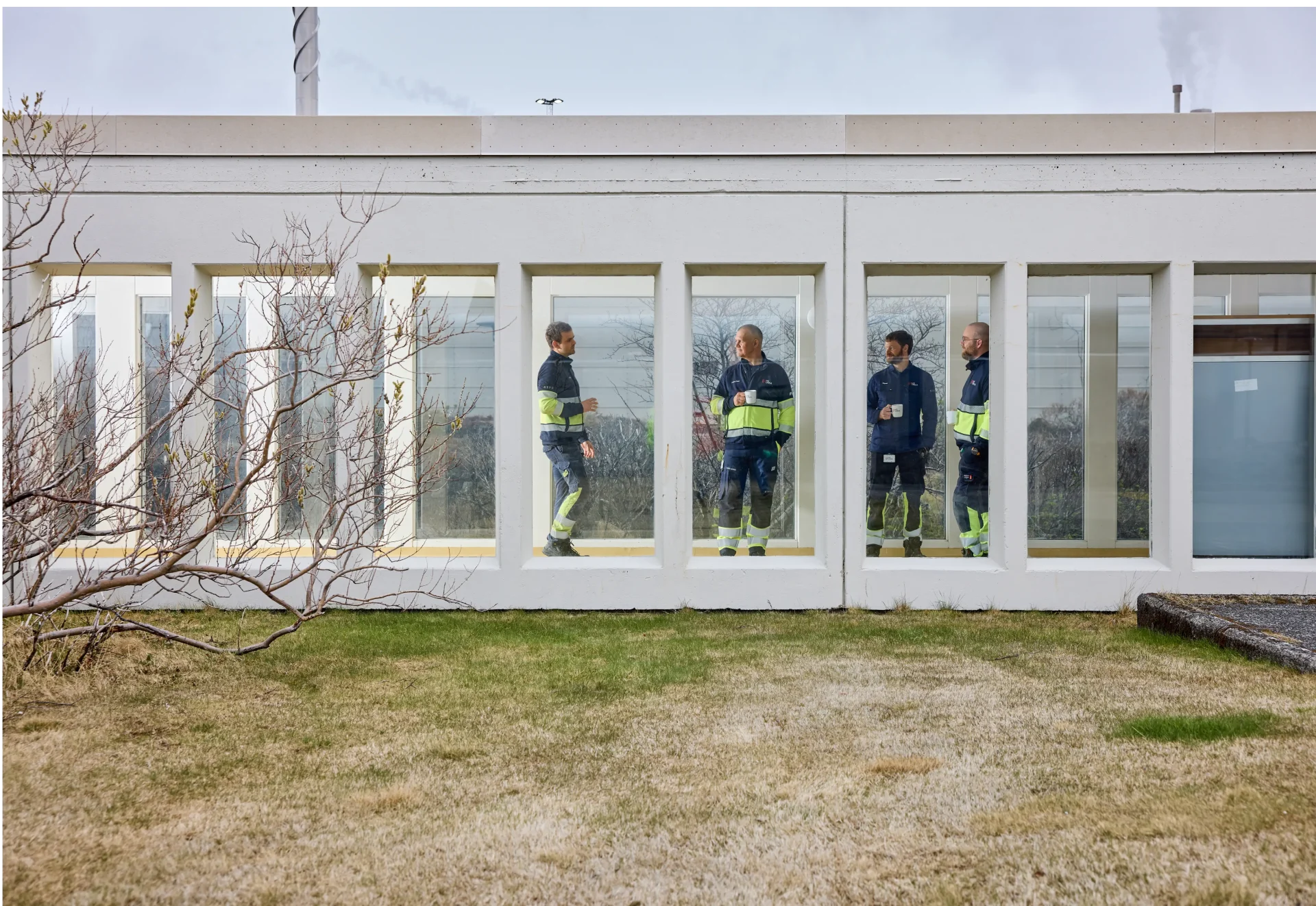 Four HS Orka employees in navy and high-visibility workwear stand talking and holding coffee mugs behind the windows of a white industrial building in Iceland, with bare trees and grass in the foreground.