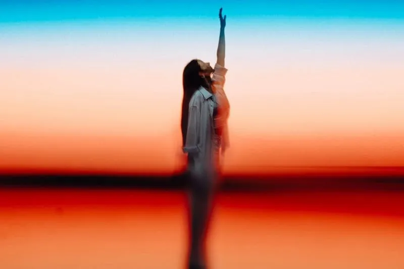 Abstract motion-blurred image of a woman in a white shirt reaching upward against a vivid blue, orange and red gradient background.