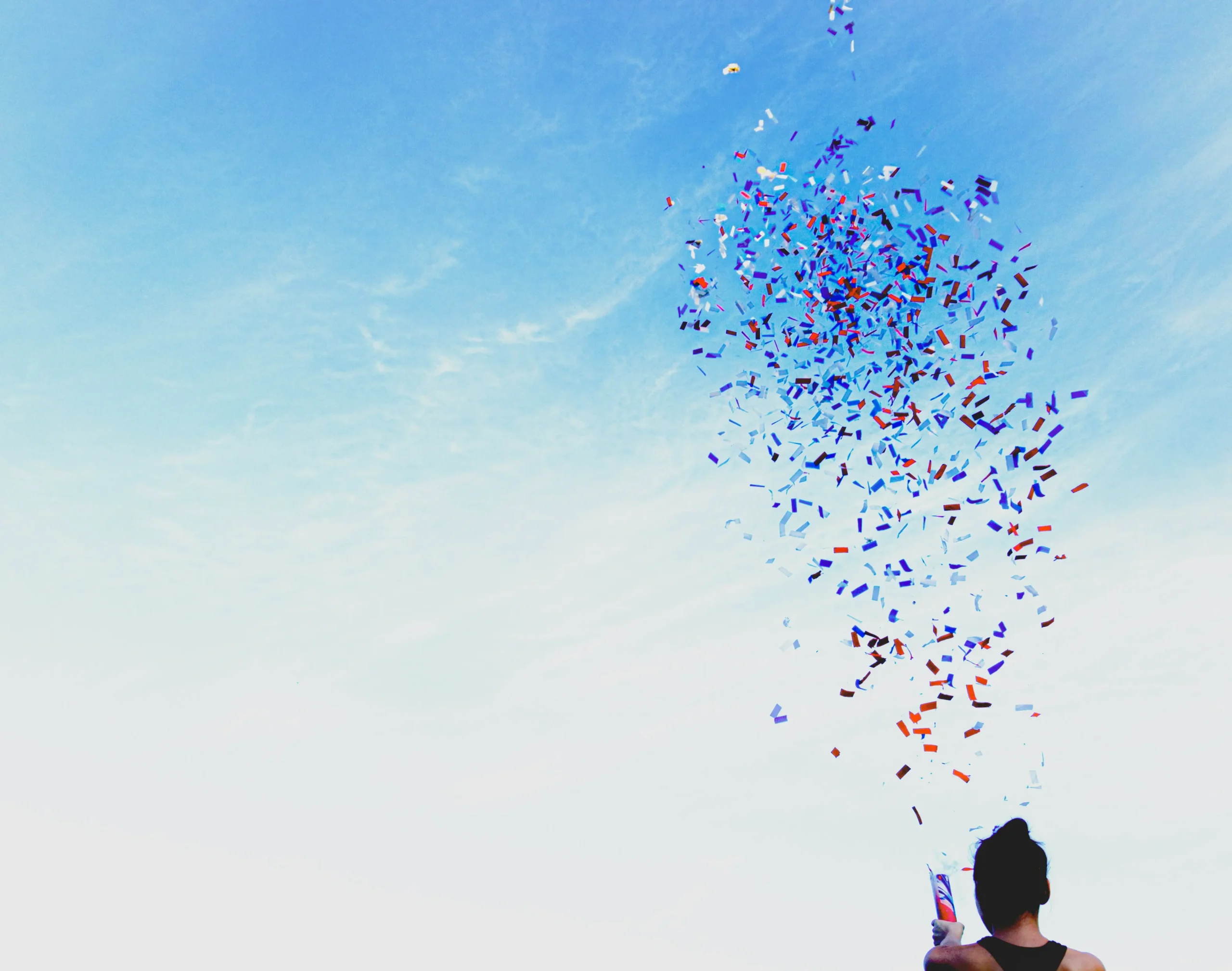 Person firing a confetti cannon into a bright blue sky, with colorful confetti swirling overhead.